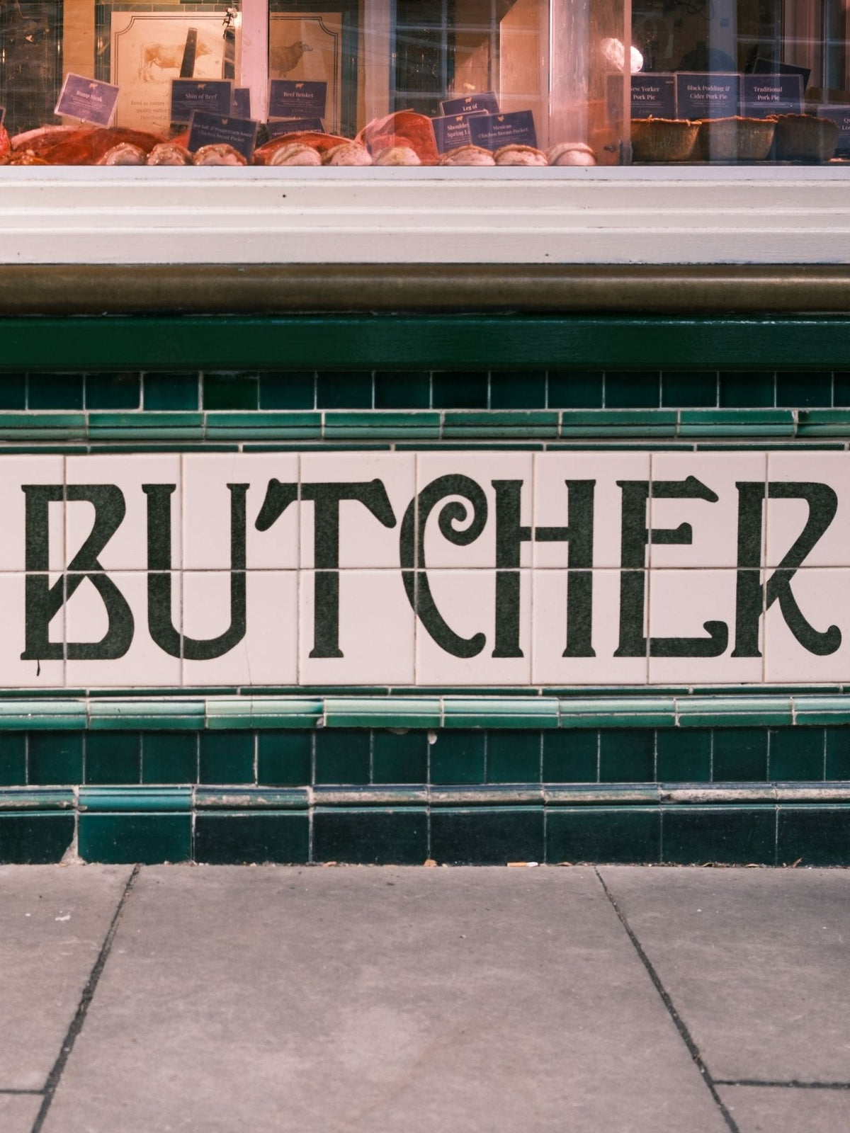 A tiled sign reading “BUTCHER” with a cuts of meat in a store window.