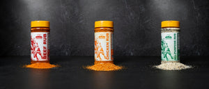 Three shakers of meat rubs lined up with piles of spice blend in front of them.