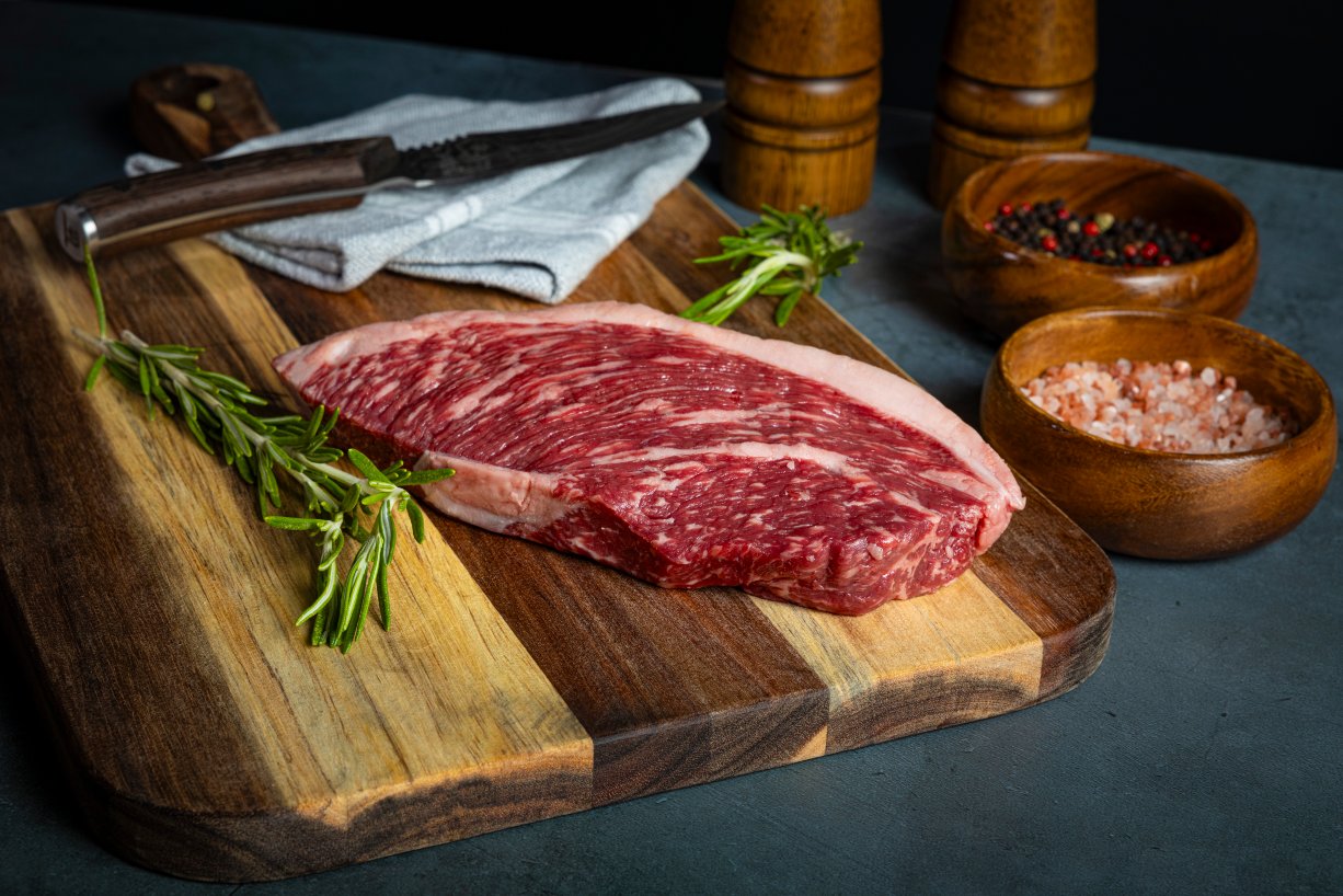 raw steak on a wooden cutting board alongside a bowl of salt, pepper, and herbs