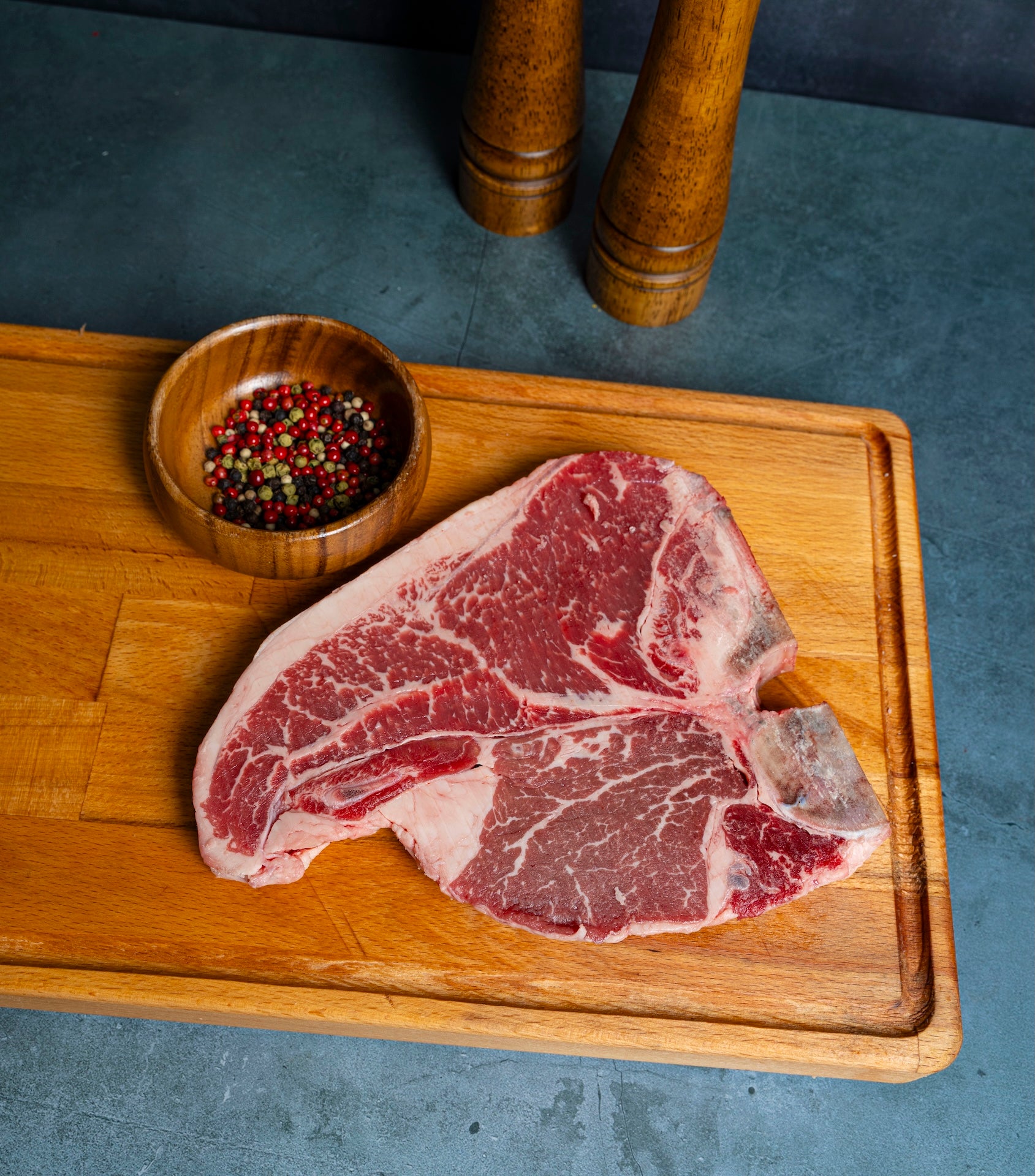 Raw porterhouse steak on a wooden cutting board with a small bowl of spices on a gray surface.