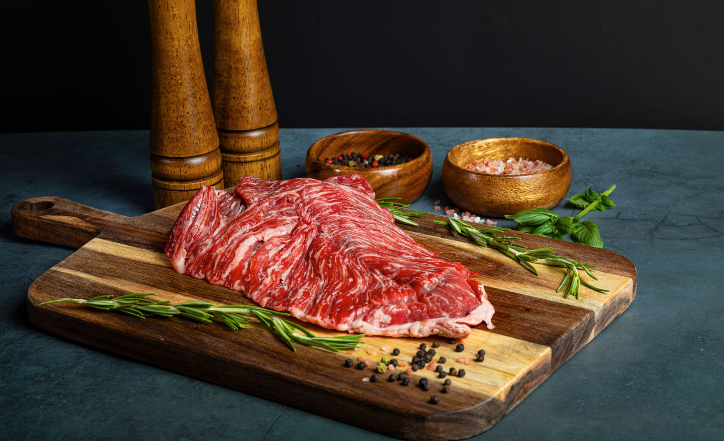 Raw Prime Sirloin Flap Meat (Ranchera) on a wooden cutting board with herbs and spices, against a dark background.