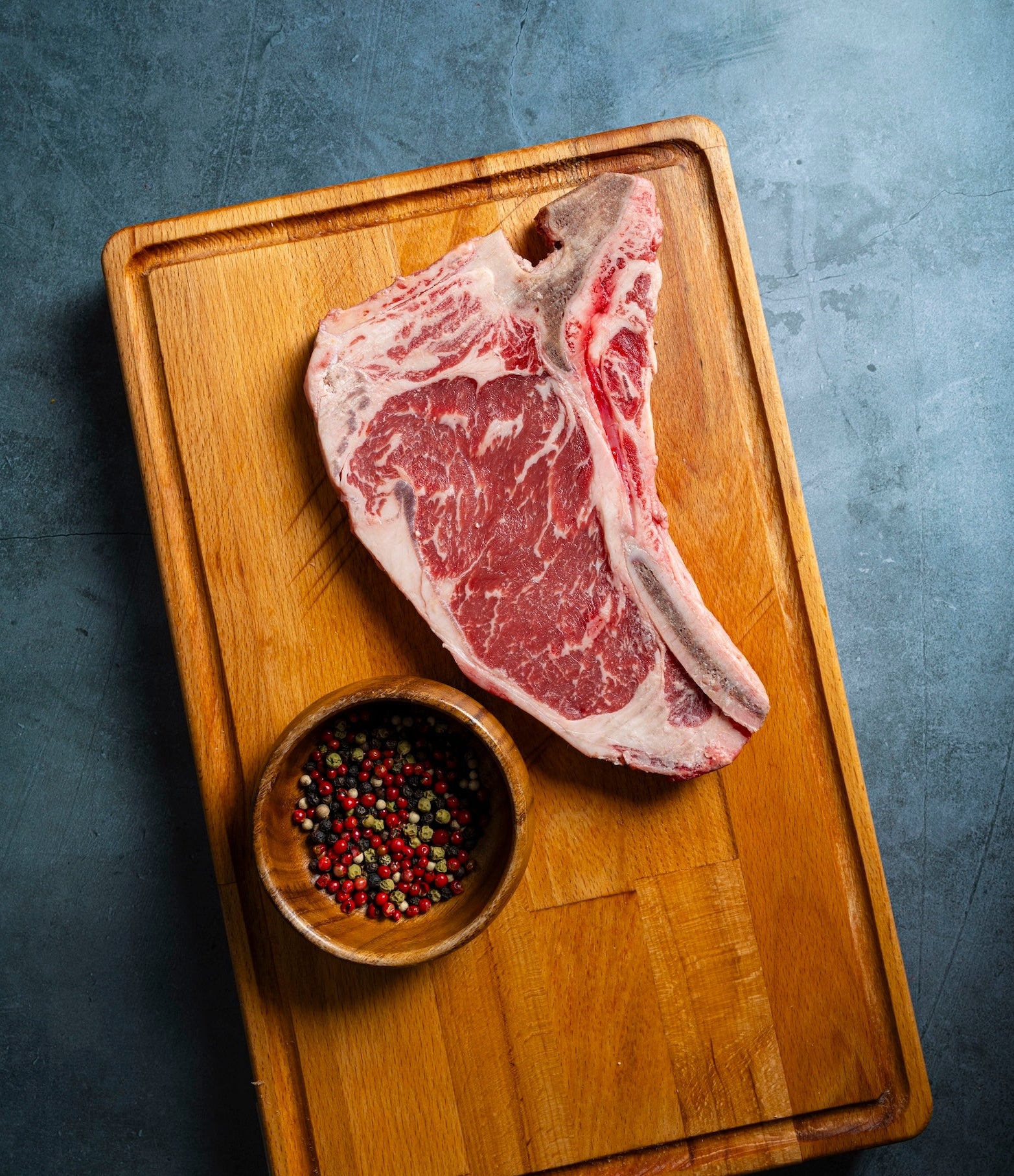 Raw steak on a wooden cutting board with a small bowl of peppercorns on a gray background