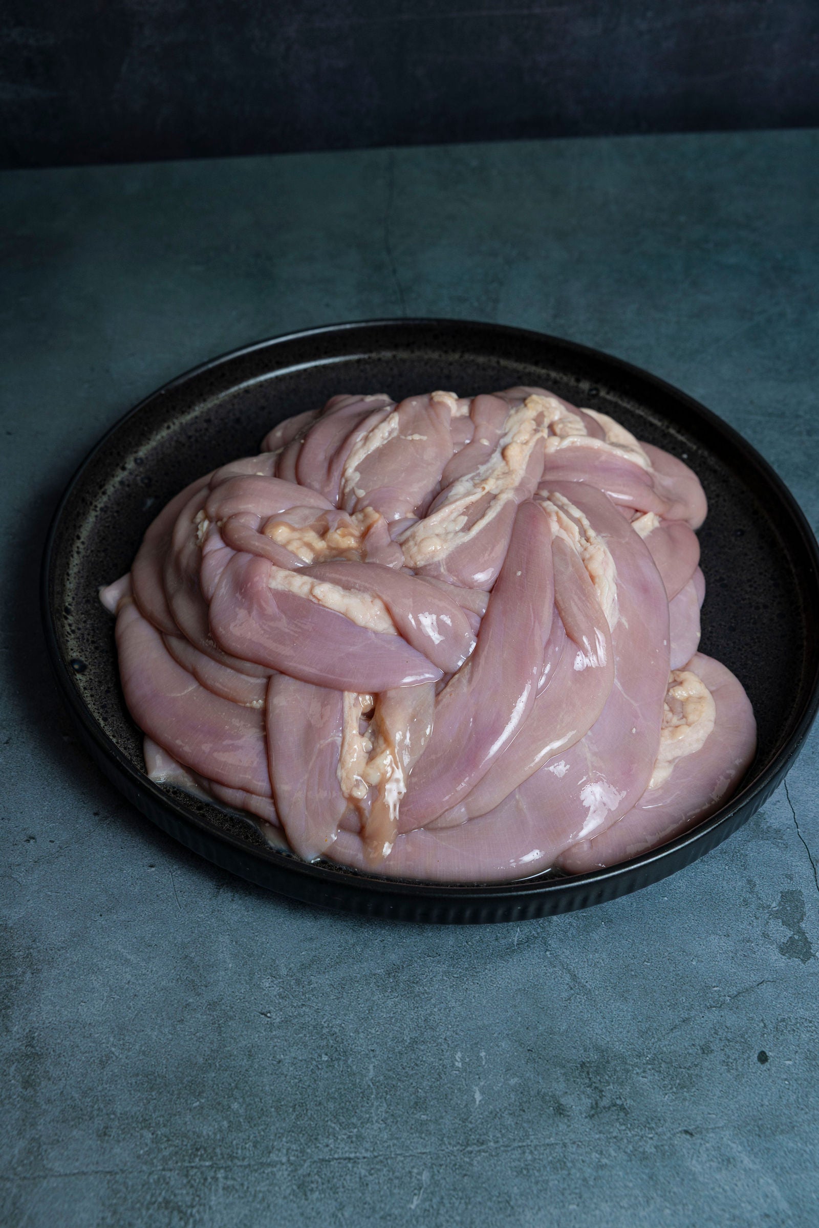 Raw chicken breast slices arranged in a braided pattern on a black plate against a dark background.
